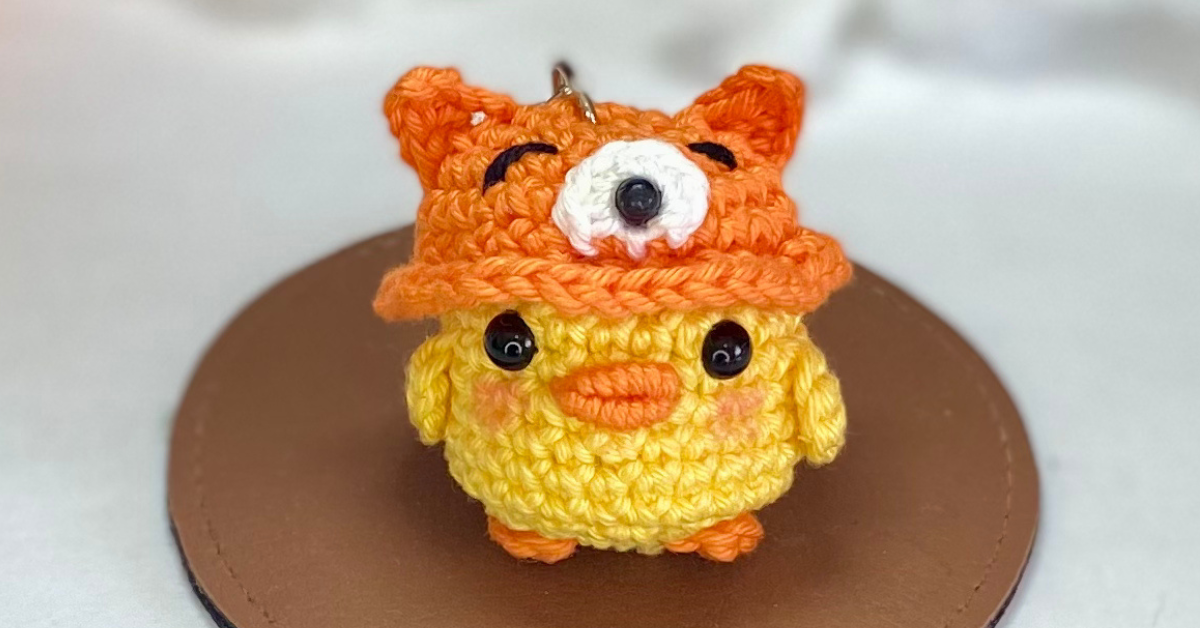 Pato amigurumi em crochê com chapéu de raposa laranja, ideal como chaveiro ou lembrancinha artesanal, ilustrando receita fácil passo a passo.