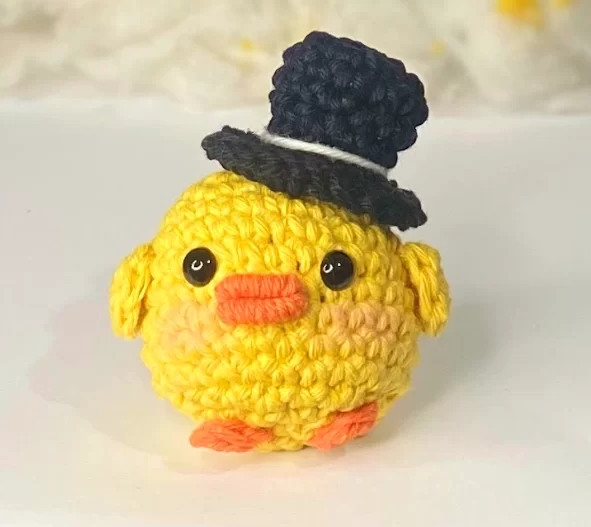 Pato amigurumi com cartola feito em crochê, com cores vibrantes e fundo claro, ideal para ilustrar o resultado final da receita passo a passo completa.