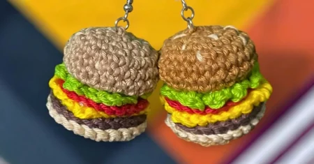 Chaveiro Mini Hambúrguer Amigurumi, Receita Grátis Passo a Passo. Par de chaveiros artesanais em forma de hambúrgueres feitos em crochê, pendurados sobre um fundo colorido com padrões geométricos em tons de amarelo, laranja, azul e listras roxas e brancas.